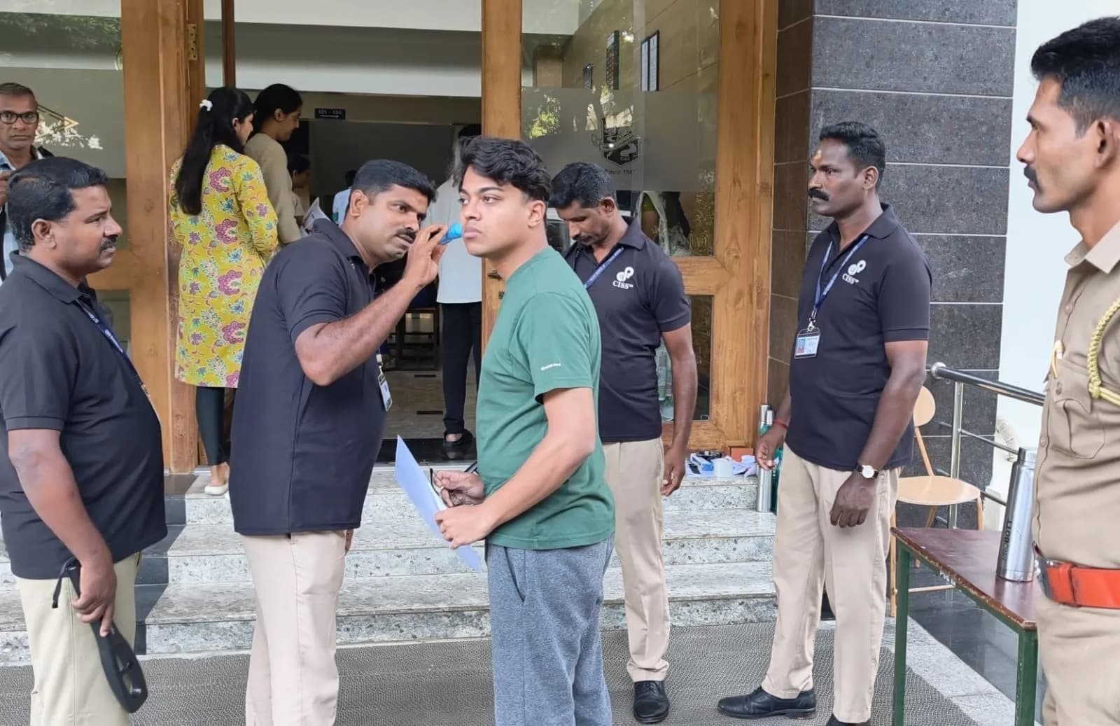 CISS personnel screening a candidate at the entrance to a NEET examination centre, with police support alongside.